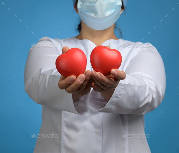 Medic in a white coat, a mask stands and holds a red heart on a blue background