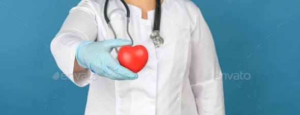 Female medic in a white coat stands and holds a red heart on a blue background