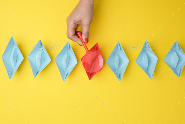 Female hand holds a magnifying glass over a row of paper boats on a yellow background