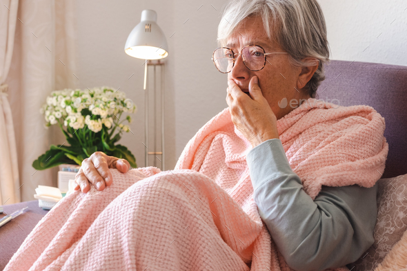 Suffering elderly woman with cough and fever as seasonal flu or pollen allergy sitting on sofa