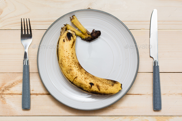 Stop wasting food. Rotten banana on a plate and cutlery on a wooden background. Food waste concept