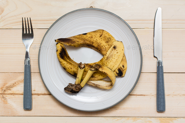 Stop wasting food. Rotten banana peel on a plate and cutlery on a wooden background.