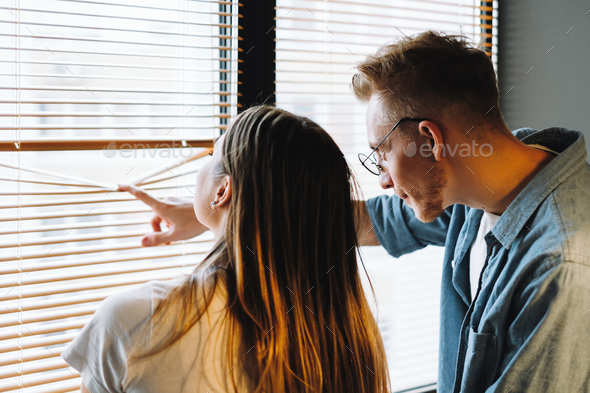 Young cheerful couple looks out the window through the blinds, looks at what is happening outdoor.