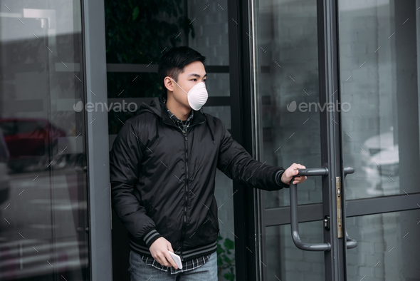 young asian man in respirator mask holding smartphone and looking away while leaving building