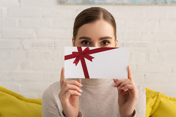 Young woman obscured face with gift card and looking at camera on sofa