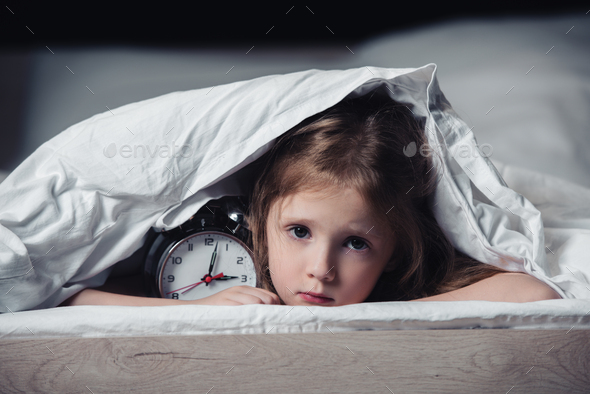 scared kid hiding under blanket with alarm clock and looking at camera isolated on black