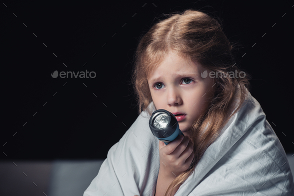 scared kid sitting under blanket and holding flashlight isolated on black