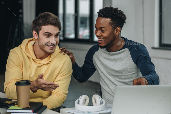 happy young programmer pointing at computer monitor while sitting near african american colleague in