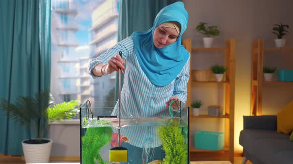 Muslim Woman in National Headscarf Cleans Aquarium with Sponge alt