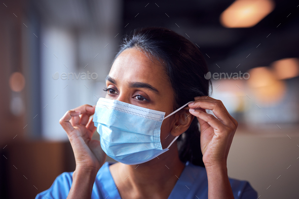 Female Doctor In Scrubs Putting On Face Mask Under Pressure In Busy Hospital During Health Pandemic
