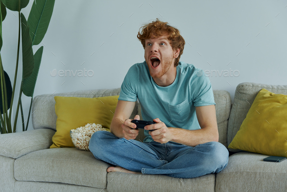 Cheerful redhead man using controller while playing video games on the couch at home