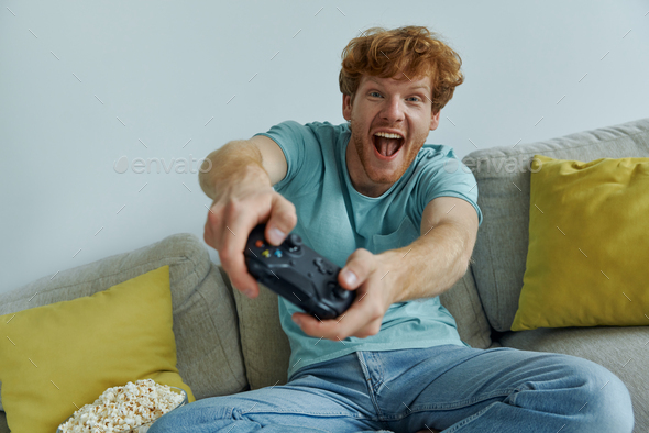 Cheerful redhead man using controller while playing video games on the couch at home