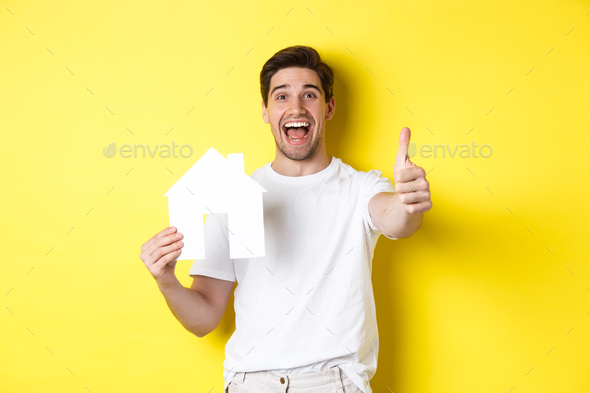 Real estate concept. Happy young male buyer showing thumb up and paper house model, smiling