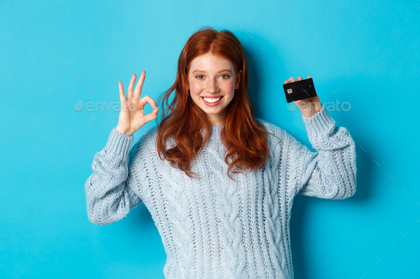 Happy redhead girl in sweater showing credit card and okay sign, recommending bank offer, standing