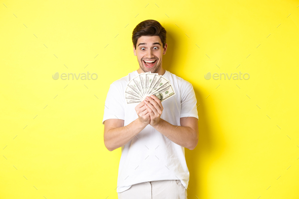 Happy man looking at money and smiling excited, winning prize, got bank loan, standing over yellow