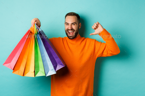Handsome bearded man in orange sweater, buying gifts, pointing at himself and showing shopping bags