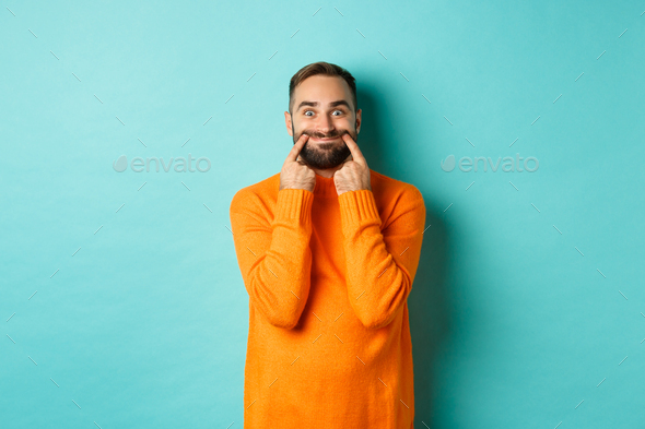 Image of bearded man stretching lips in happy smile, faking happiness, standing over light blue