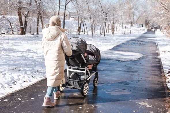 Young mother with double baby stroller for twins walking outside in winter