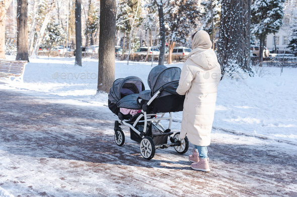 Young mother pushing a baby stroller for twins walking outside in winter