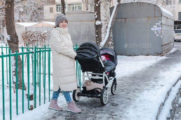 Young mother with double baby stroller for twins walking outside in winter