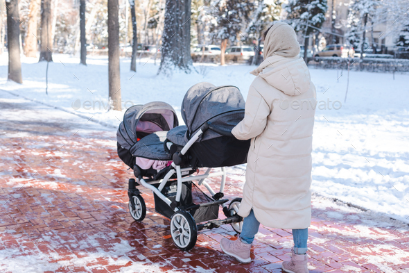 Young mother pushing a baby stroller for twins walking outside in winter