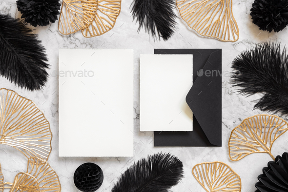 Cards and envelope on marble table near black feathers and golden leaves. Wedding mockup