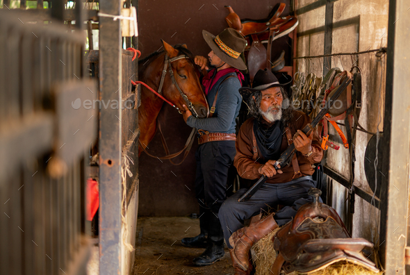 Two man with cowboy costume stay in horse stable that senior check long gun