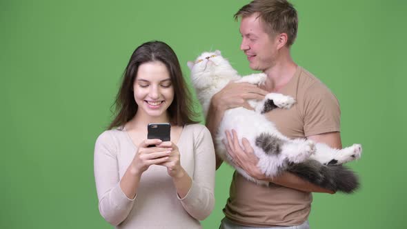 Young Couple Using Phone Together with Cat Against Green Background alt