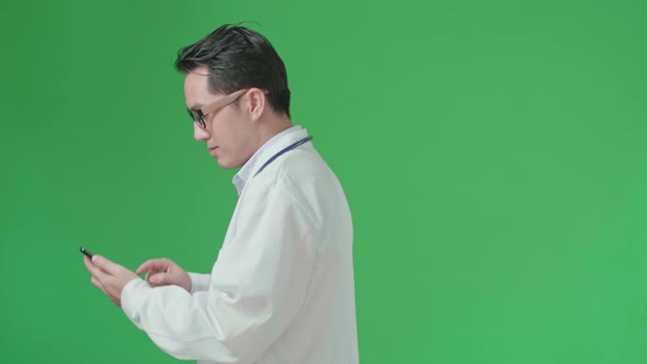 Side View Of Asian Doctor With Stethoscope Using Mobile Phone While Walking On Green Screen alt