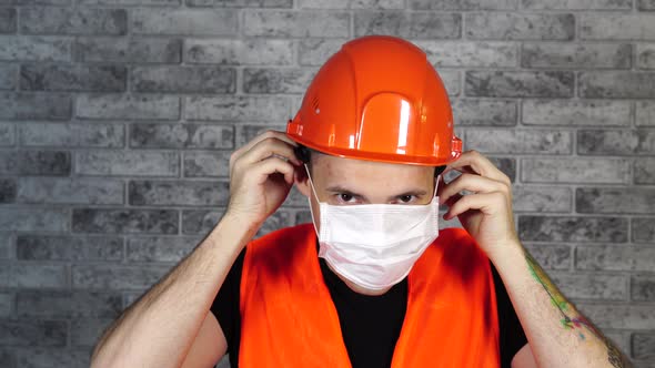 Male Construction Worker in Overalls Putting on Medical Mask on Face on Background of Gray Brick alt