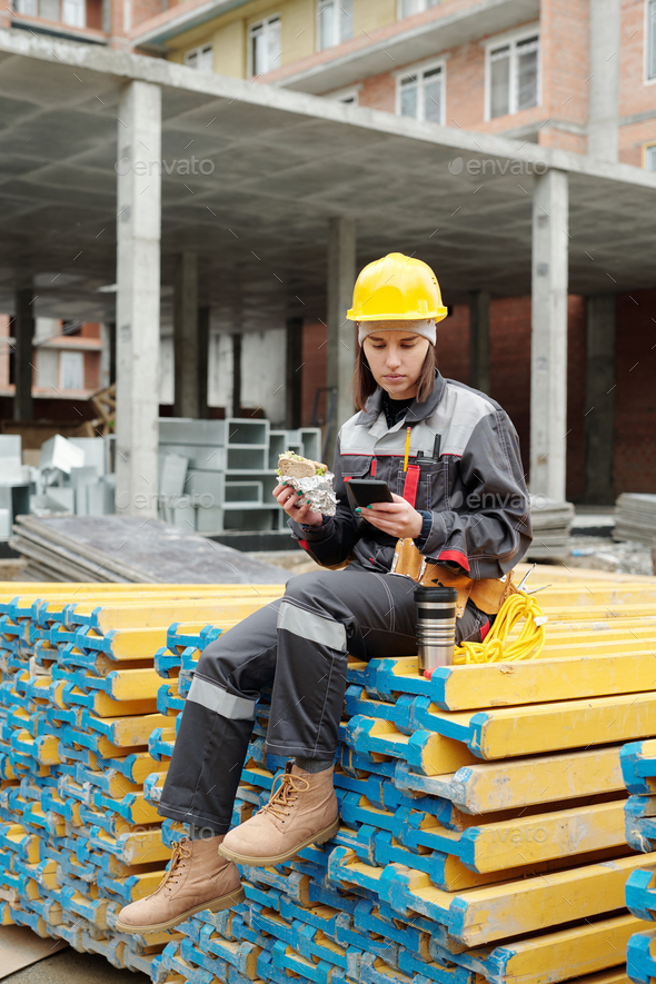 Young female builder in workwear having sandwich and using smartphone