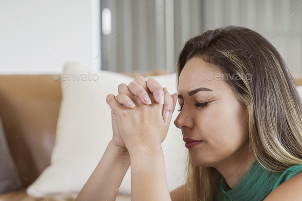 Hispanic woman praying with eyes closed at home, online church concept
