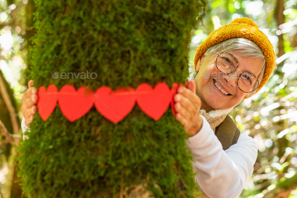 Smiling elderly woman hugging a moss covered tree trunk in the woods with red heart shaped papers