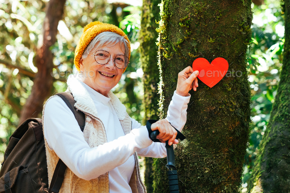 Smiling elderly woman hiking in the woods holding a paper heart near a moss covered tree trunk