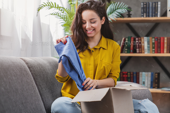 Young happy girl woman customer sit on sofa unpack parcel delivery box with clothes