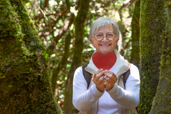 Smiling elderly woman holding a paper heart near a moss covered tree trunk in the woods