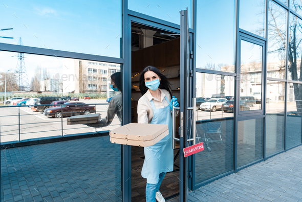 Cafe owner in medical mask near door with card with quarantine lettering showing boxes and looking