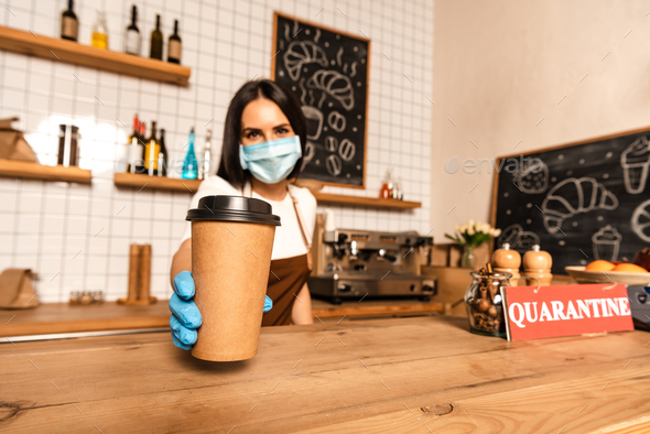 Selective focus of cafe owner in medical mask showing paper cup of coffee near table with card with