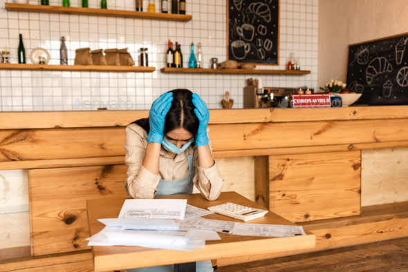 Stressed cafe owner in latex gloves touching head near documents and calculator at table