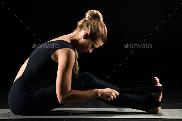 Side view of young woman practicing yoga and stretching with yoga strap isolated on black