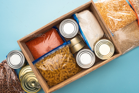 top view of cans and groats in zipper bags in wooden box on blue background, food donation concept