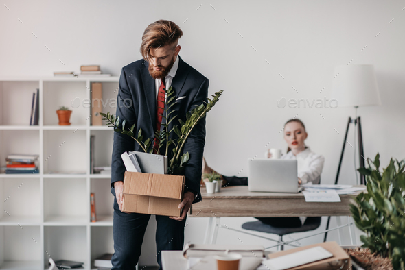 Young upset fired businessman with cardboard box in office, boss behind