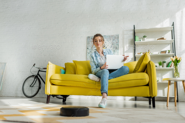 attractive woman using laptop while robotic vacuum cleaner washing carpet in living room