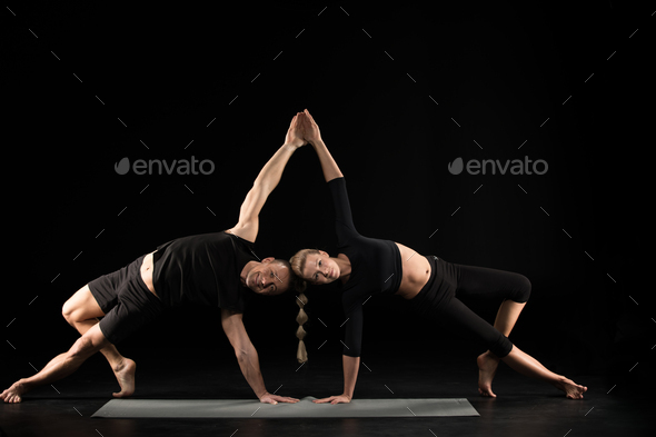 Couple performing acroyoga side plank pose on yoga mat isolated on black