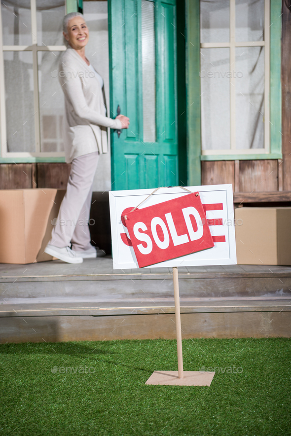 Smiling senior woman standing on porch of new house with sold sign