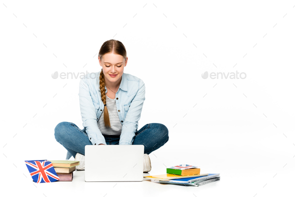 smiling girl sitting on floor using laptop near books and copybooks, uk flag isolated on white