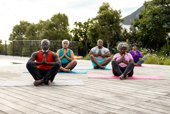 Multiracial senior friends meditating on mats while sitting on hardwood floor against plants in yard - Stock Photo - Images