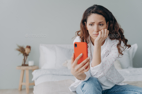 Stressed caucasian woman is doom scrolling. Girl reading morning news on smartphone in her bedroom.
