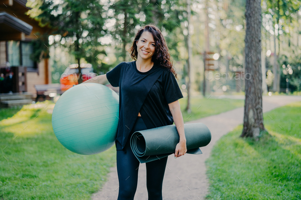 Woman smiles positively holds big fitness ball and karemat, dressed in black t shirt.