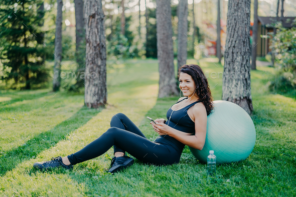 Woman takes break after aerobics exercises with fitness ball, sits on green grass.